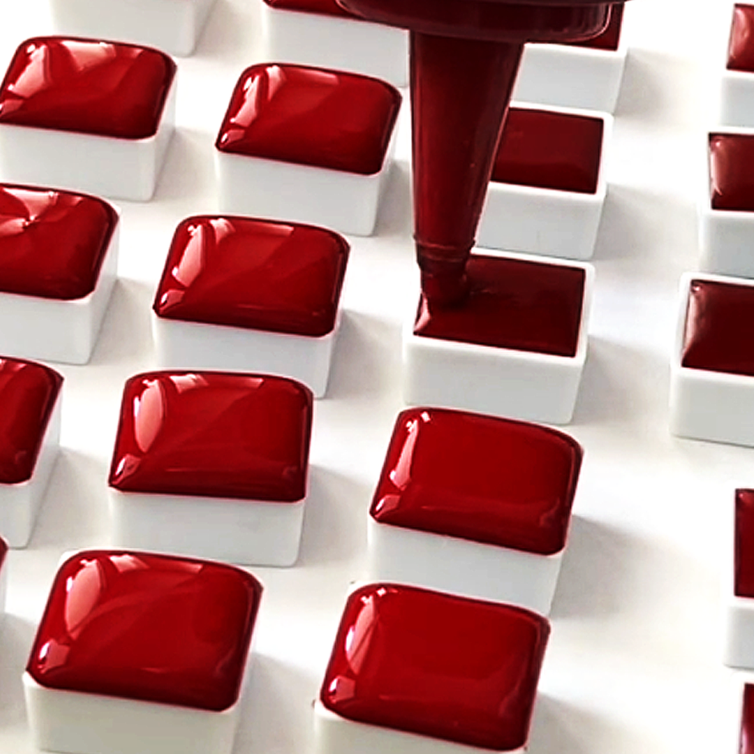 Precision dispensing nozzle filling white plastic half-pans with liquid crimson watercolor paint by VHacademy